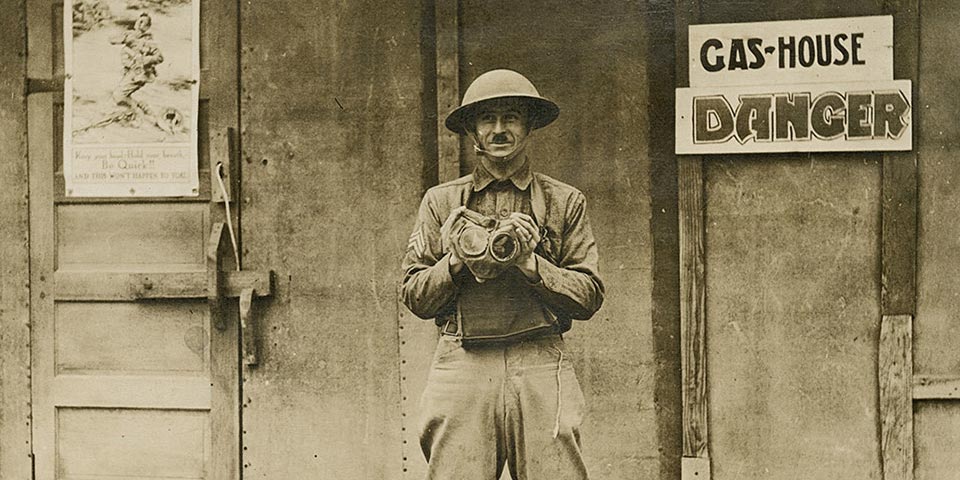 Man in uniform holding a gas mask standing in front of a building with a sign that says 'Gas House'