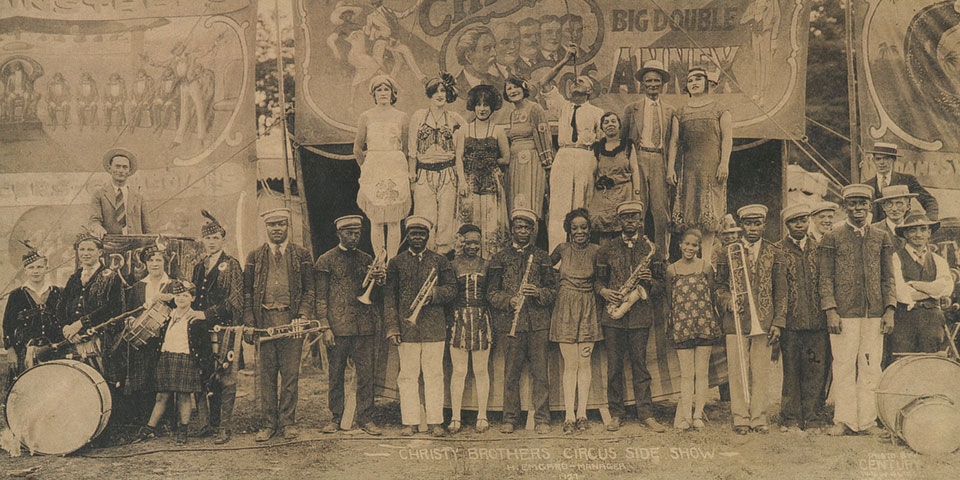 Black and white photograph of a group of sideshow performers and musicians standing in front of banners advertising the Christy Brothers Circus and various sideshow acts.