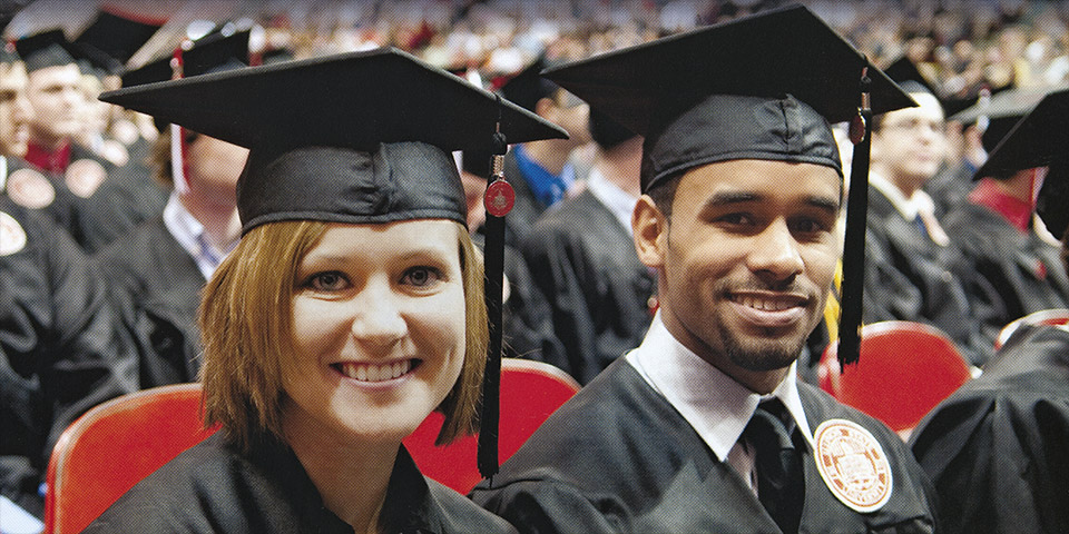 Two smiling students in front of a crowd wearing graduation cap and gown