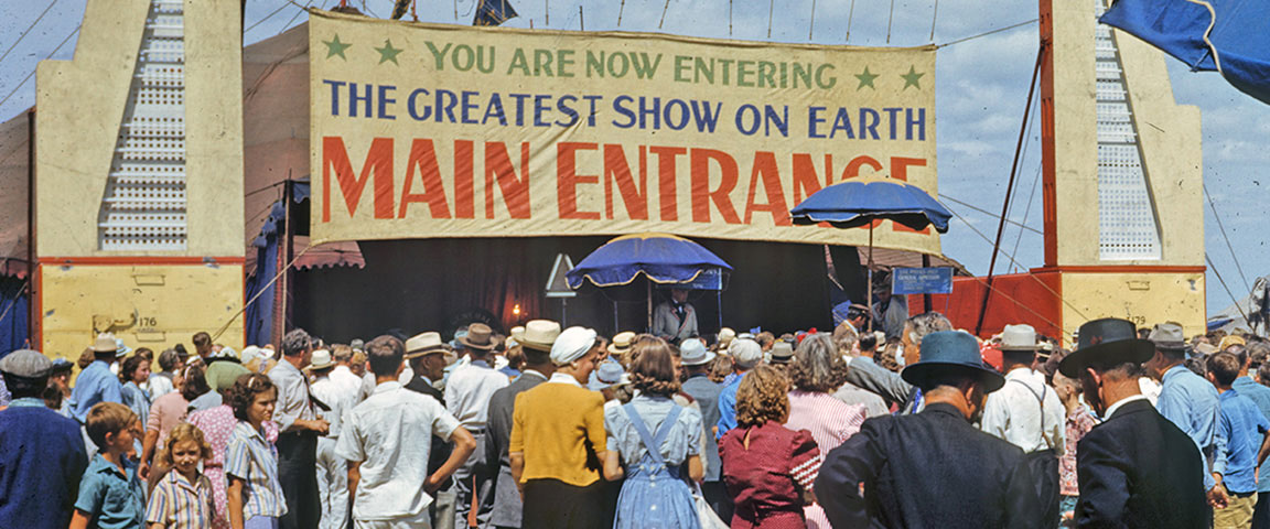 Crowd of customers moving towards a tent labeled 'You are now enering the greatest show on earth. Main Entrance.'