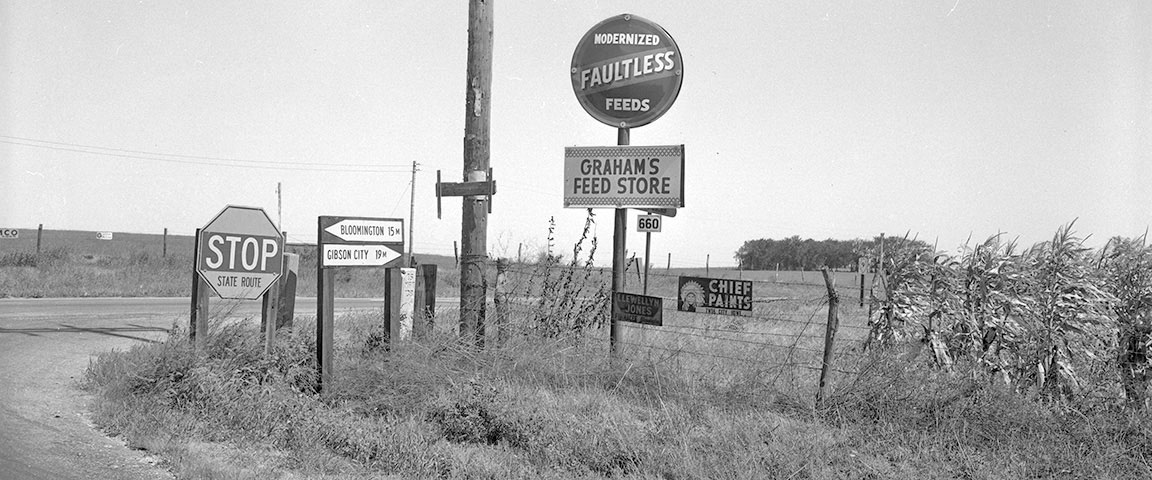 Black and white photo of signs flanking a country road next to a corn field. 